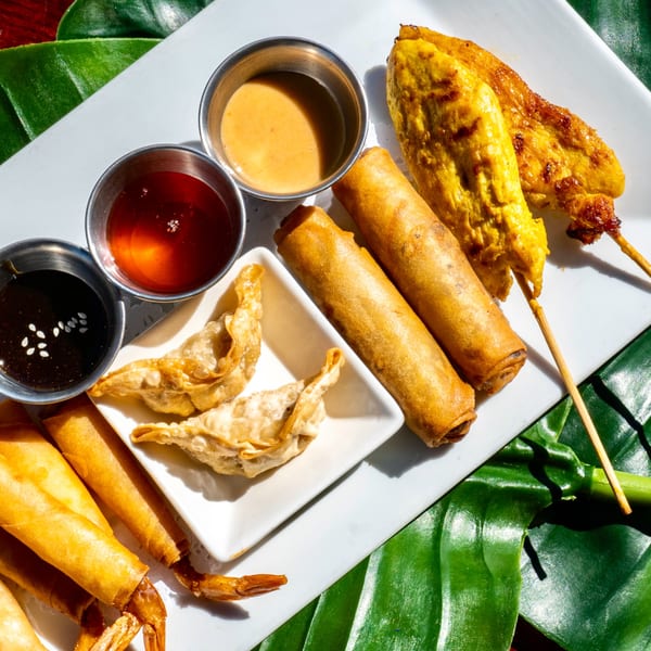A white platter of Thai appetizers including chicken satay, spring rolls, and crab rangoons with dipping sauces.