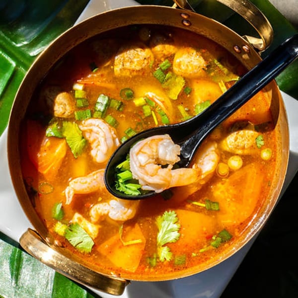 Close-up of Thailicious' Tom Yum soup with shrimp, mushrooms, and herbs in a brass pot.