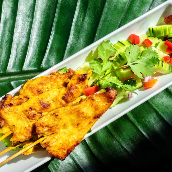 Thailicious chicken satay skewers with cucumber salad and cilantro on a banana leaf.