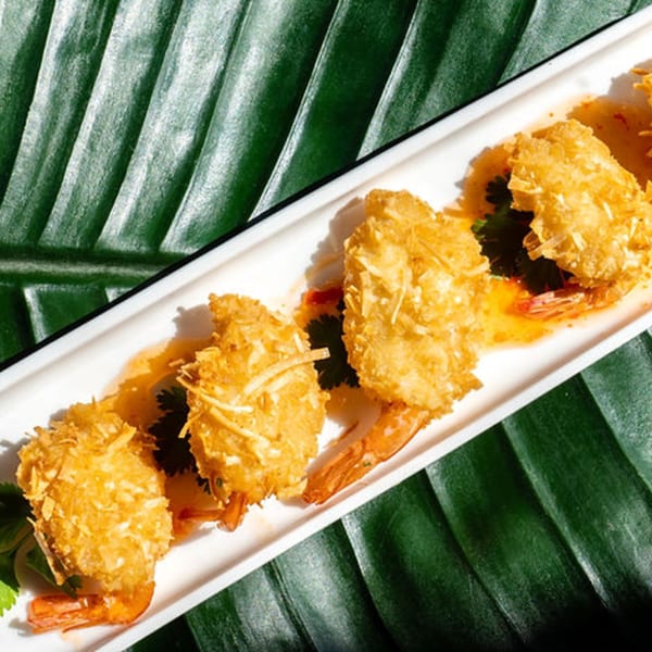 Four crispy coconut shrimp with sweet chili sauce on a white plate, against a green leaf background.