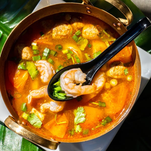 A brass pot of Thailicious Tom Yum soup with shrimp, mushrooms, and herbs, with a spoon.