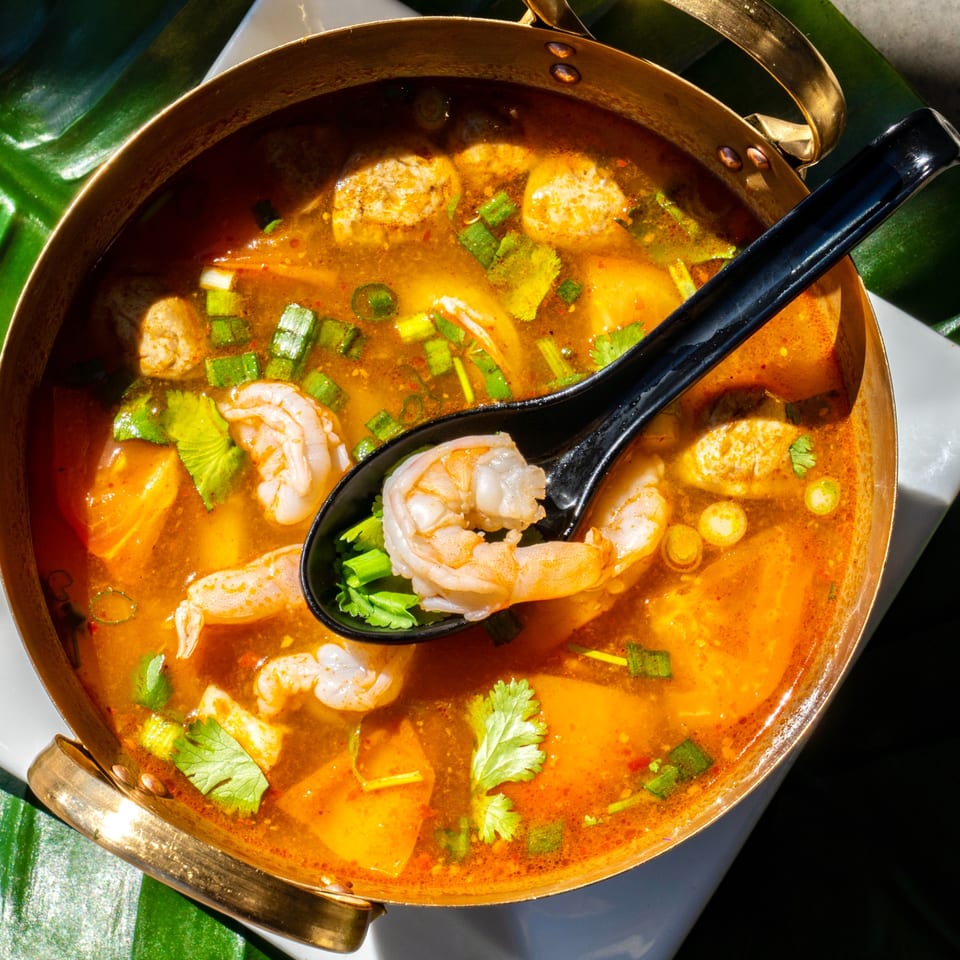 A brass pot of Thailicious Tom Yum soup with shrimp, mushrooms, and herbs, with a spoon.