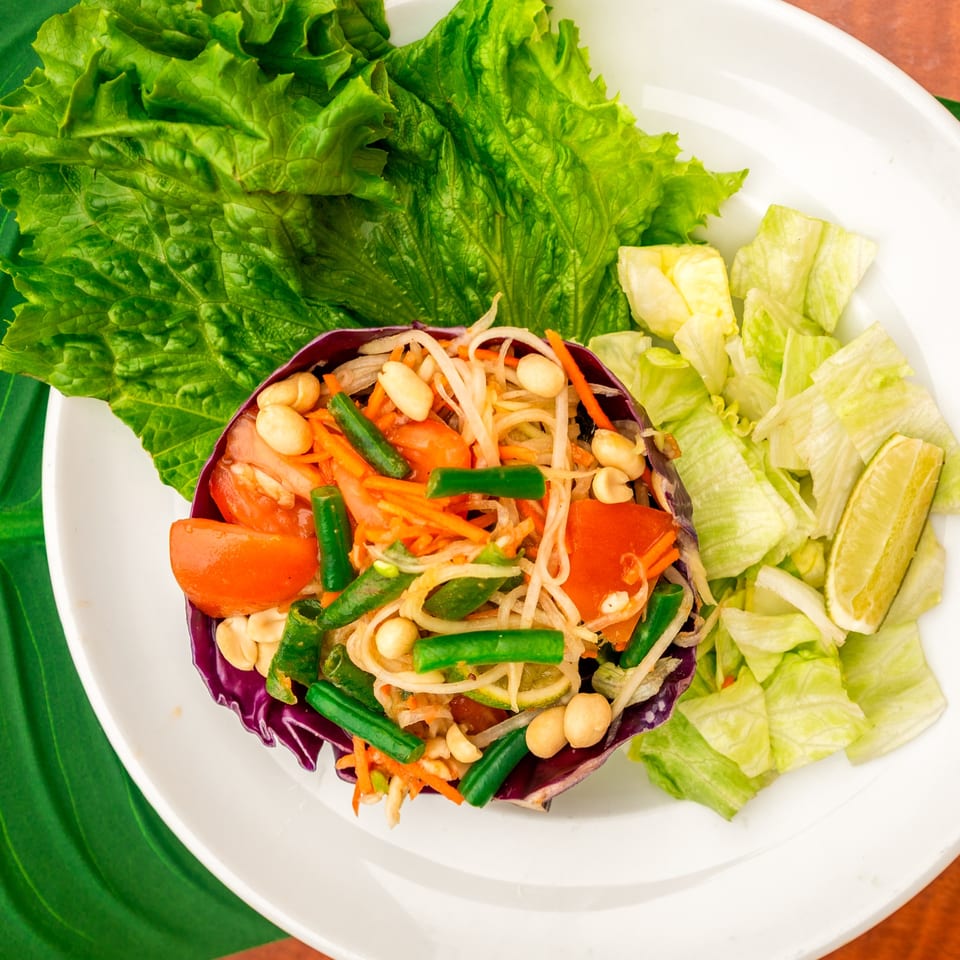 Thailicious papaya salad with green beans, tomatoes, peanuts, and lime on a white plate.