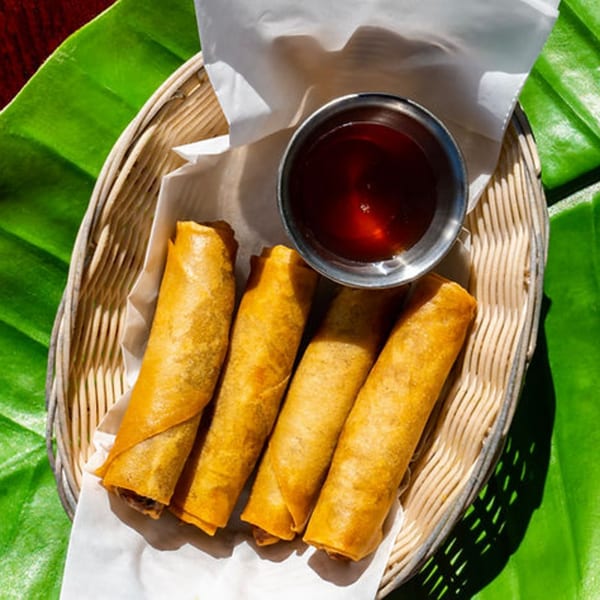 Four crispy Thailicious spring rolls in a basket with dipping sauce, on a large green leaf.